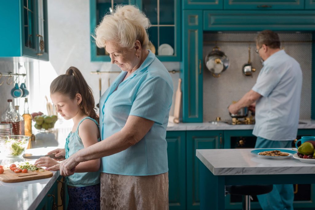 An elderly woman assists a young girl with cutting vegetables in a teal-colored kitchen, while an elderly man cooks on the stove in the background.